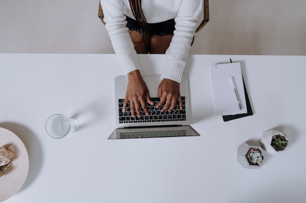 Mesa de trabalho branca com diversos objetos e computador; Moca de blusa branca com as mãos sobre o teclado. Visão do alto.
