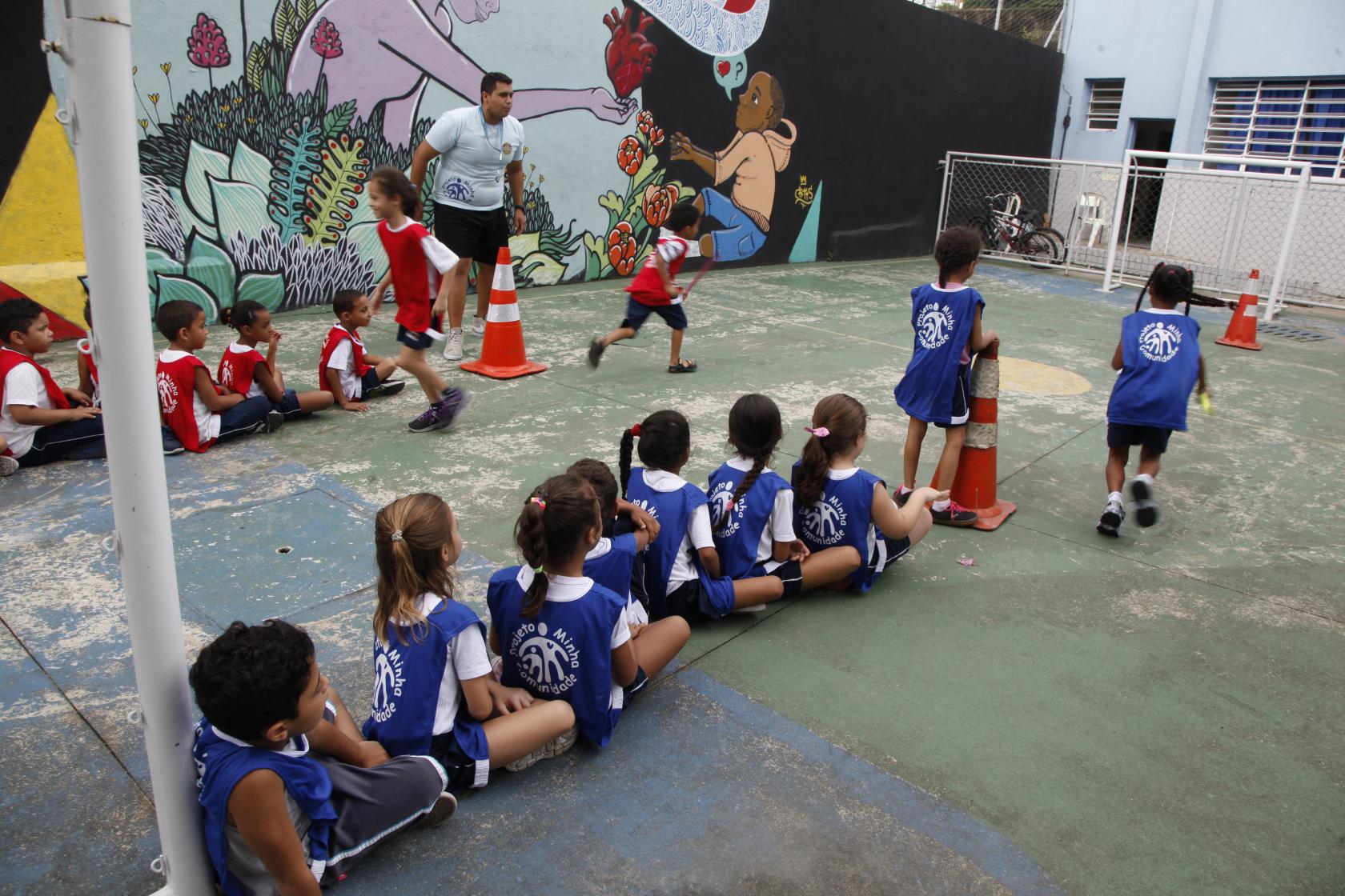 Crianças em pátio de escola pública durante atividade física, com professor ao fundo. Crianças estão distribuídas em duas filas e usam bola para a atividade.