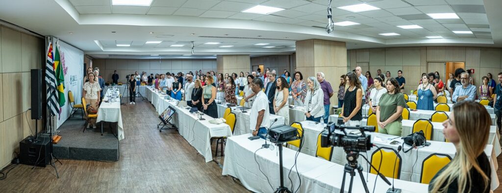 Foto da reunião dos conselheiros e conselheiras do CAU São Paulo. Mostra sala com mesas e tablado, com audiência e componentes da mesa diretora em posição para audição do Hino Nacional.