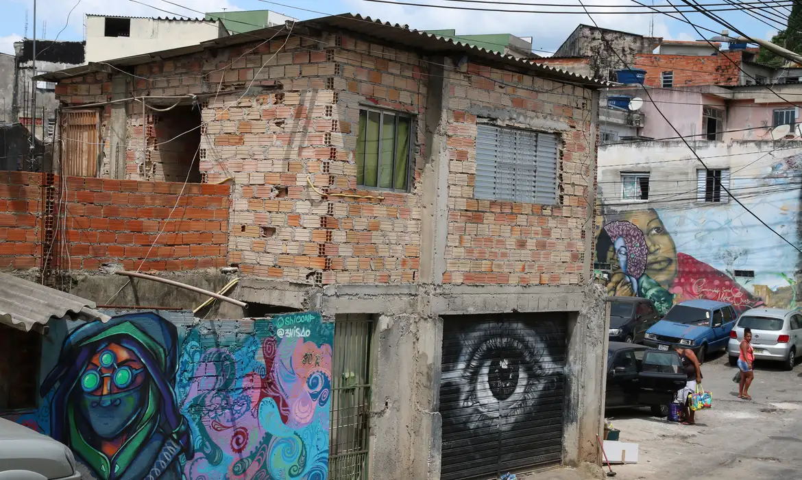 Foto mostra residencia em favela com as paredes sem cobertura e desenhos (grafites) sobrepostos a um portal de metal e um muro.