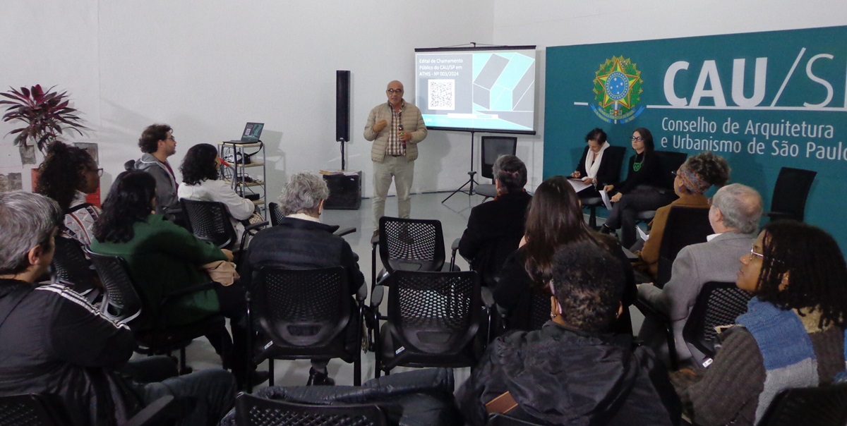 #PraCegoVer Fotografia salão com uma audiência assistindo a um palestrante, homem de óculos e de pé com um microfone, com uma ampla faixa verde ocupando quase toda a parede da direita, com o logo do CAU São Paulo em destaque.