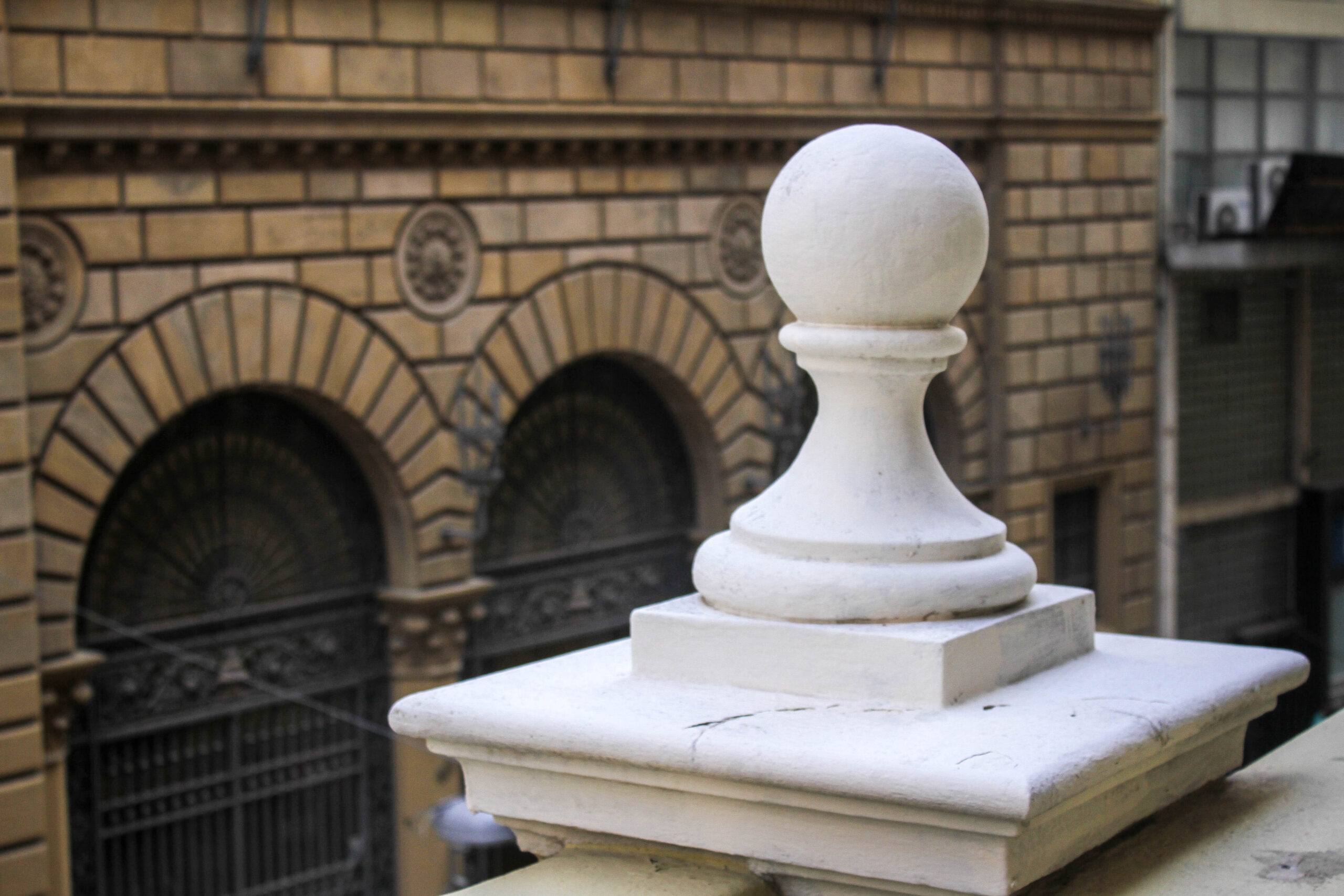 #PraCegoVerDetalhe da arquitetura do prédio da sede do CAU São Paulo. A foto é um 'close-up' de uma balaustrada de pedra branca com um pináculo esférico no topo, em frente a um edifício com detalhes arquitetônicos ornamentados, incluindo janelas arqueadas e trabalhos decorativos em pedra.