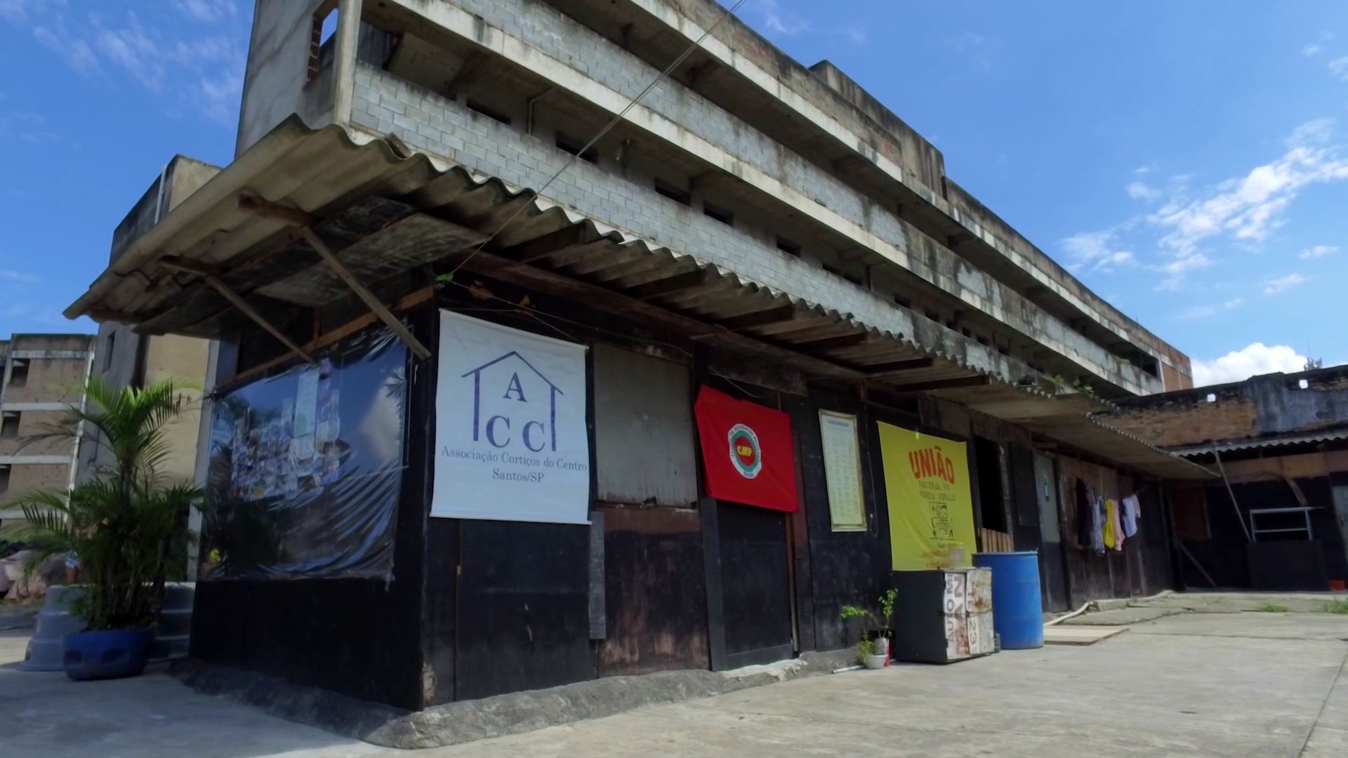 #PraCegoVerFoto reproduz uma cena do curta-metragem sobre moradia. A imagem mostra um construção de um piso com diversas placas com nomes das organizações de luta por habitação popular.