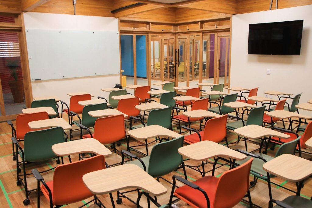 #PraCegoVerFotografia mostra o interior de um sala de aula, com assentos das cores laranja e verde, com uma tela branca ao fundo e uma televisão na parede lateral.
