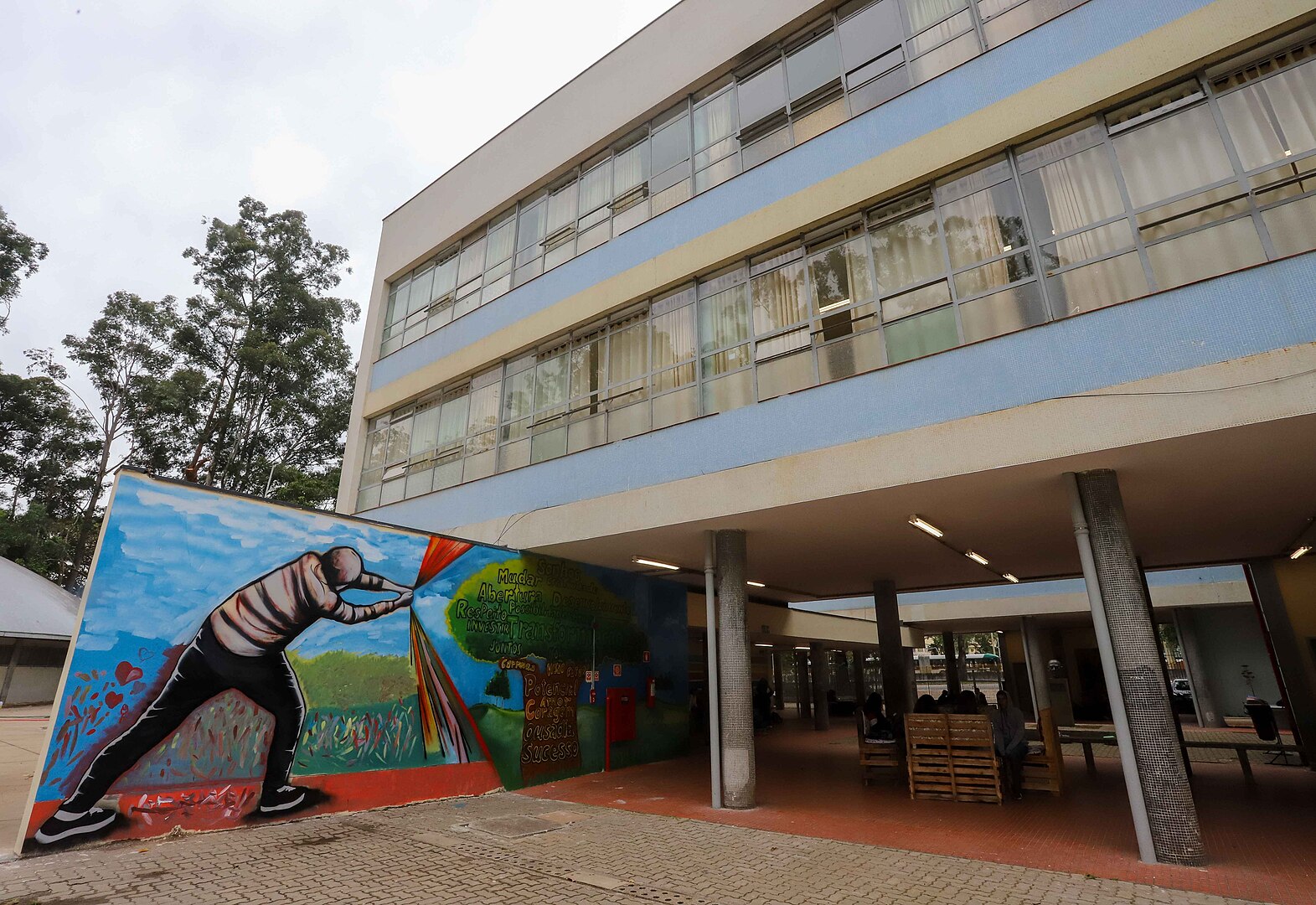 #PraCegoVerFotografia mostra parte de um prédio público, apoiado sobre pilotis, com algumas poucas pessoas em um espaço de convivência. Em destaque, há um muro com um grafite colorido. 