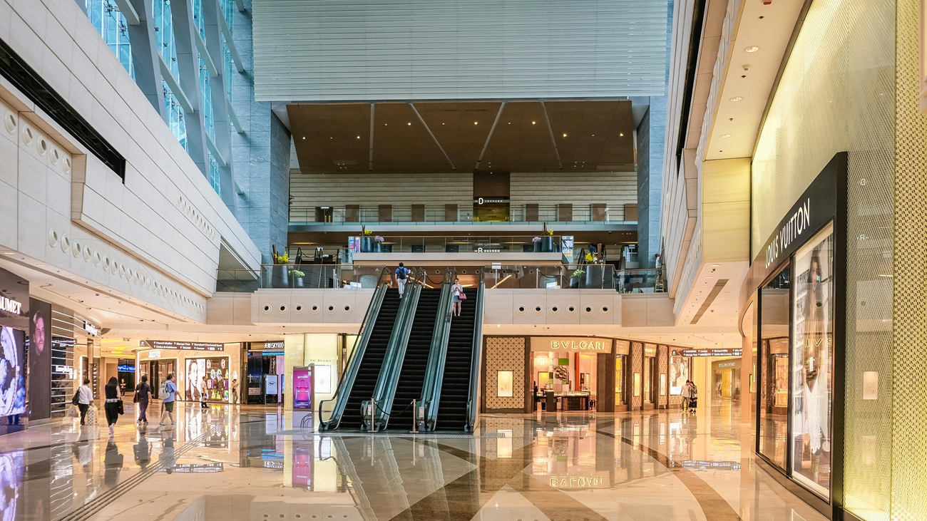 #PraCego#Ver Fotografia mostra imagem do interior de um shopping center, com três escadas rolantes ao centro e várias lojas. Alguns poucos usuários passeiam pelos corredores.