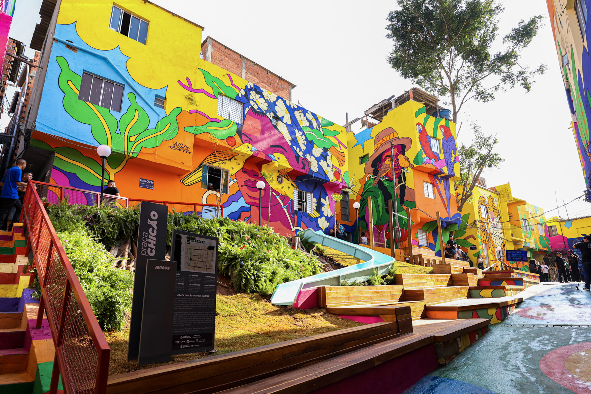 #PraCegoVerA fotografia mostra uma cena vibrante de arte urbana. Há prédios com paredes coloridas, apresentando grandes murais de folhas verdes e outros motivos de plantas. No primeiro plano, há bancos de madeira e degraus pintados em tons de amarelo e laranja, ao lado de uma fonte com azulejos azuis. Este espaço público parece ser projetado tanto para apelo estético quanto para engajamento comunitário, combinando arte, arquitetura e função social. (Texto gerado por IA, com edição humana).