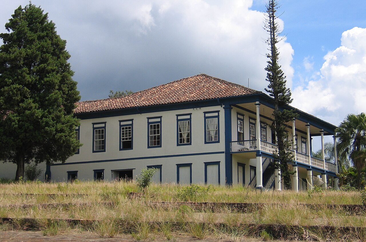 #PraCegoVerA fotografia mostra um edifício de dois andares com um estilo arquitetônico tradicional, apresentando paredes brancas e detalhes em azul ao redor das janelas e cantos. 
O telhado é de telhas vermelhas e há uma escada externa que leva a uma varanda no andar superior, que envolve a estrutura. Em frente ao edifício, há uma área gramada e descuidada com degraus que levam até ele, e há árvores altas, incluindo pinheiros, ao redor do local. 
