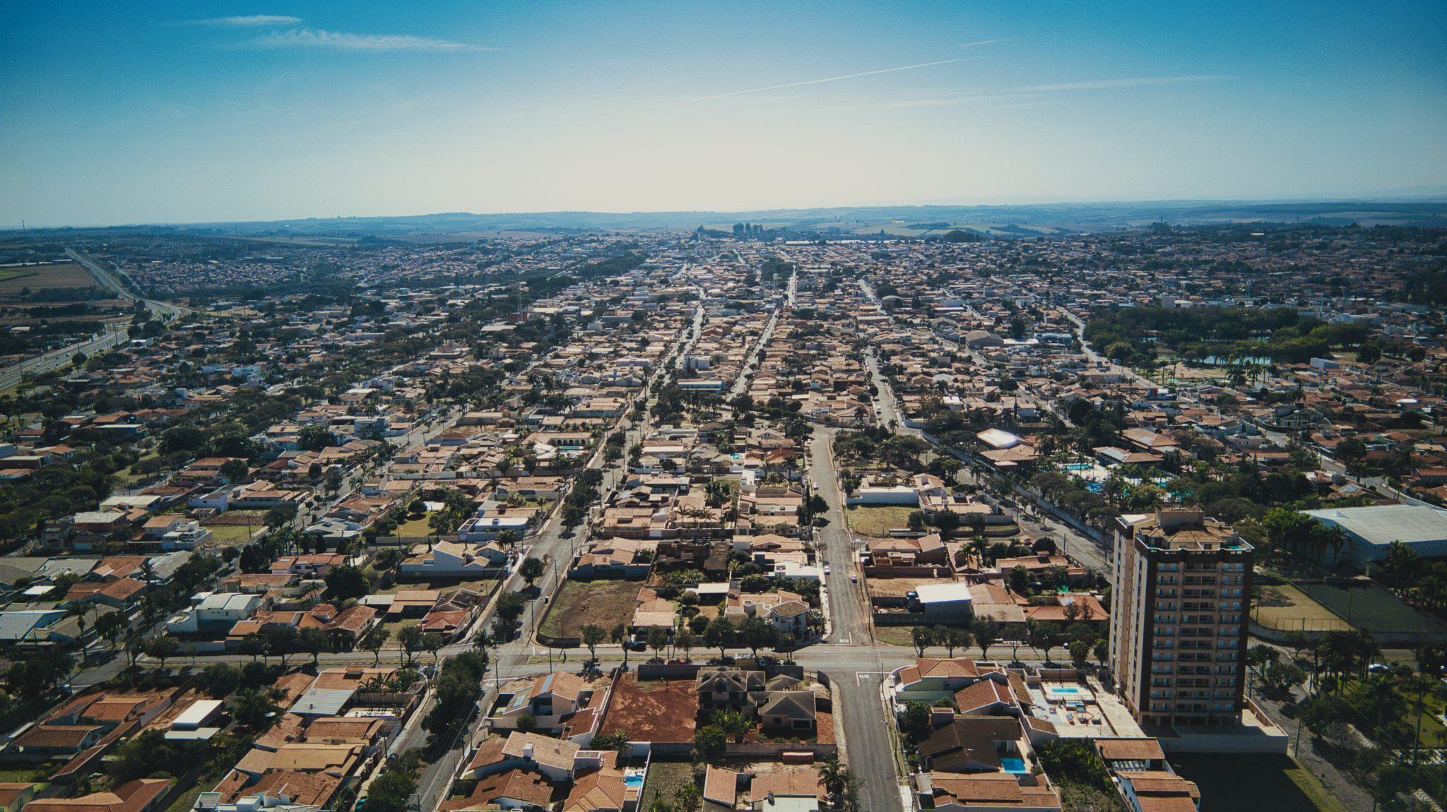 #PraCegoVerVisão do alto do município de Artur Nogueira. A fotografia mostra uma visão ampla da cidade, com uma maioria de edificações baixas e um prédio de 12 andares no canto direito da imagem.