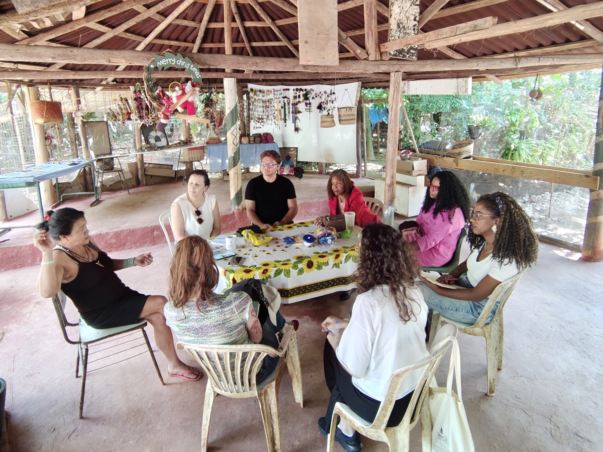 #PraCegoVer Fotografia mostra grupo de pessoas em torno de uma mesa pela manhã. O ambiente é aberto, com uma cobertura de pau a pique sustentada por toras de madeira.