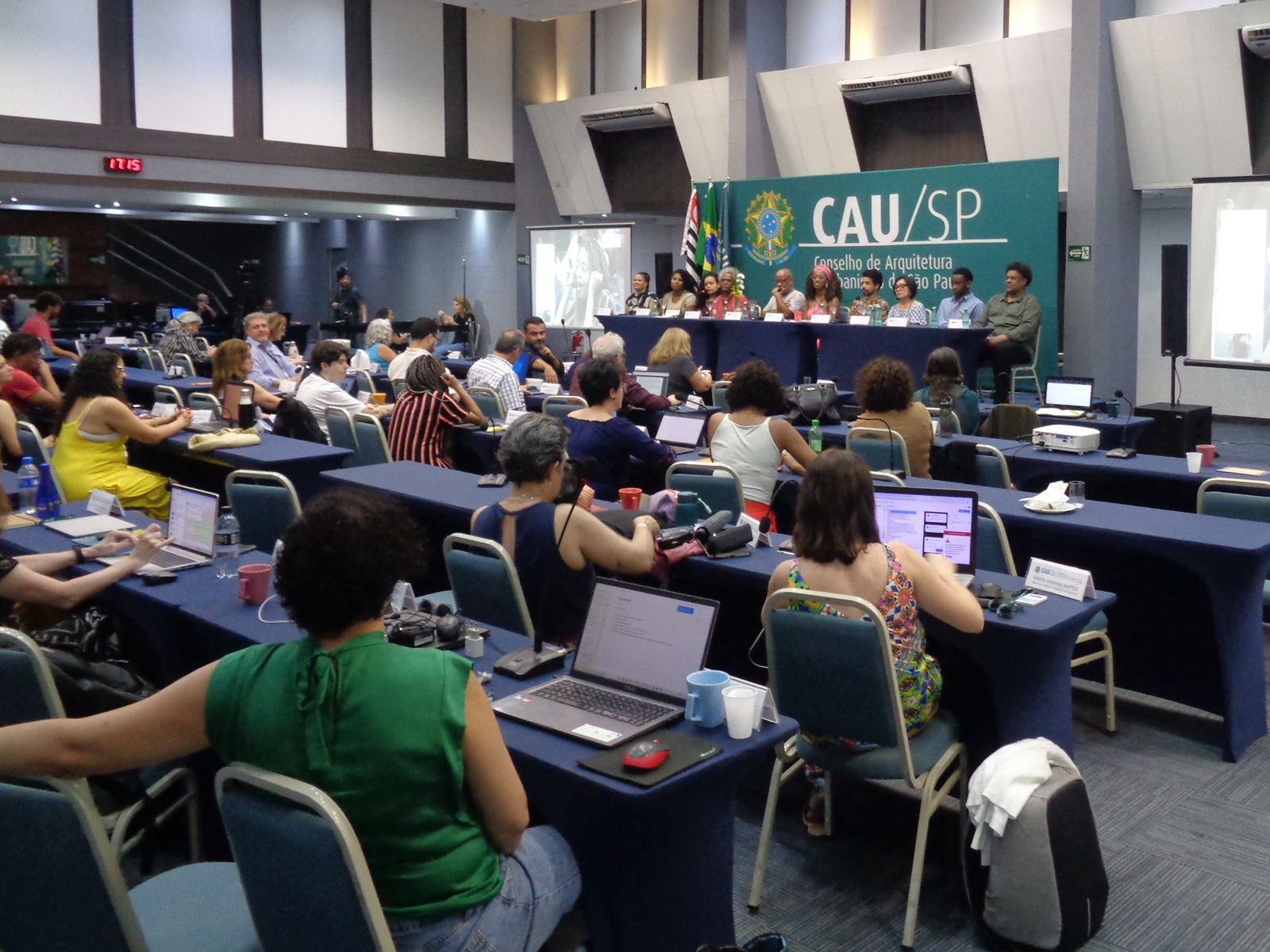 #PraCegoVerAudiência fotografada de costas observa mesa com 10 pessoas num tablado. Atrás da mesa há a tradicional tela verde com o logotipo do CAU São Paulo e bandeiras à esquerda. Dois telões à esquerda e à direita guarnecem a mesa.