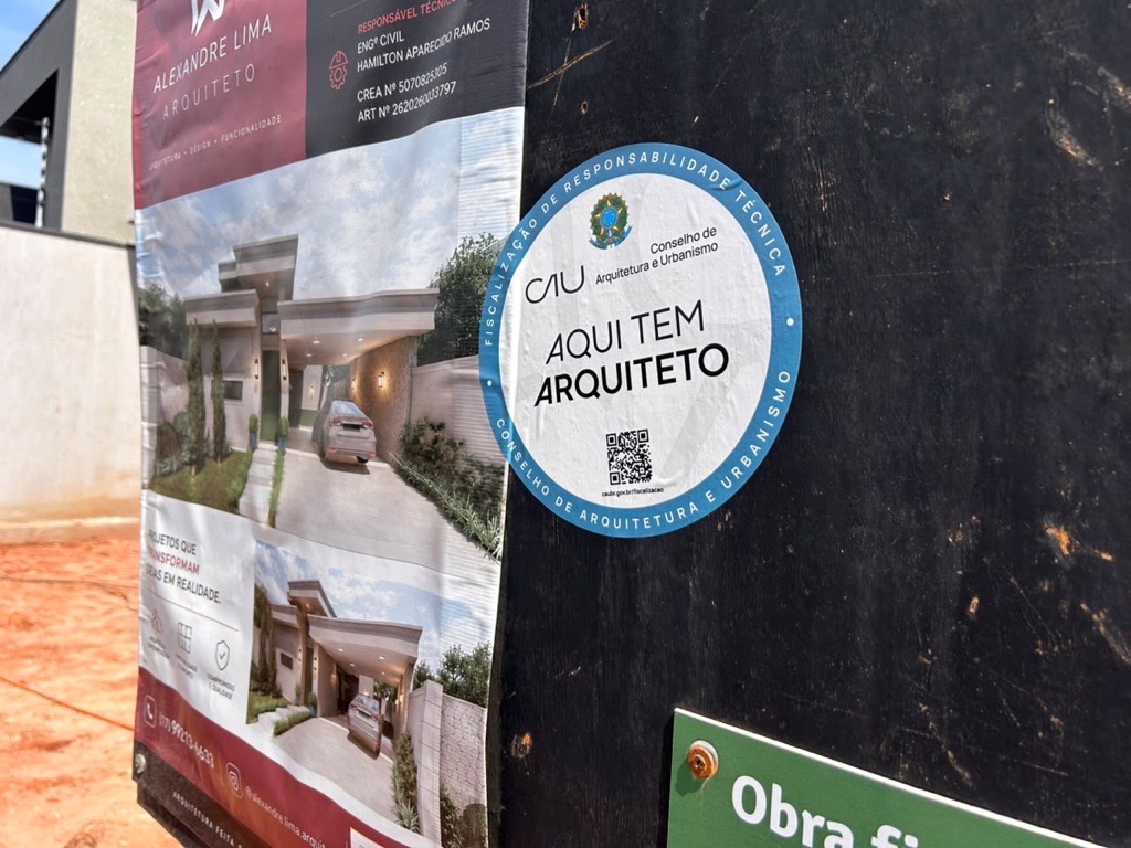 #paratodosverem
A imagem mostra um canteiro de obras com um banner exibindo o projeto de uma casa moderna — fachada com linhas retas, garagem e jardim. Ao lado, há um selo circular do Conselho de Arquitetura e Urbanismo (CAU) com os dizeres “Aqui tem arquiteto” e um QR code, indicando a responsabilidade técnica do profissional.
Descrição feita por IA.
