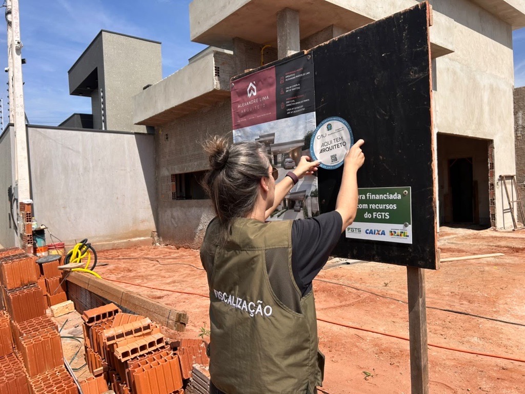 #paratodosverem
A imagem mostra uma mulher com colete verde escrito “FISCALIZAÇÃO” nas costas, em frente a uma obra em andamento. Ela aplica sobre um painel preto o selo circular do CAU com os dizeres “Aqui tem arquiteto”. Ao fundo, vê-se uma construção moderna de concreto e pilhas de tijolos sobre o chão avermelhado.
Descrição feita por IA.
